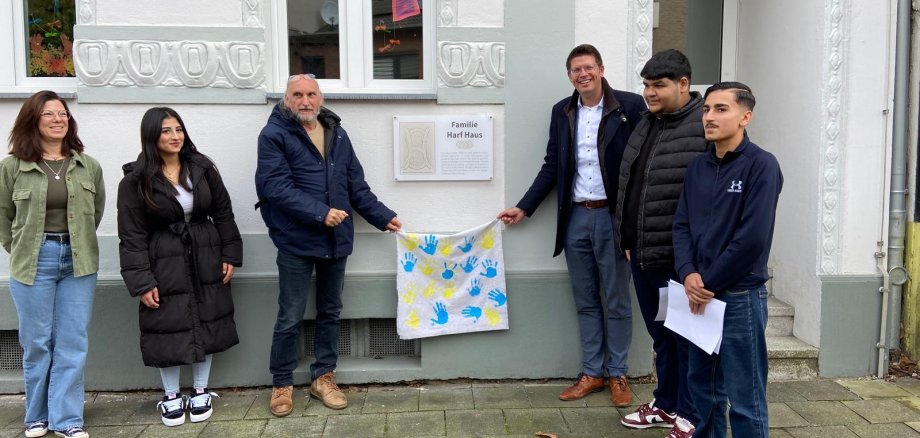 Kita-Leiterin Eva Jabs (l.), Hubert Rütten vom Heimatverein (3.v.l.) und Bürgermeister Stephan Muckel (3.v.r.) enthüllen gemeinsam mit Schüler*innen der Gemeinschaftshauptschule das neue Schild am "Familie Harf Haus".